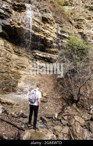 Ragazzo biondo con zaino grigio vicino alla cascata. Foto verticale. Foto Stock