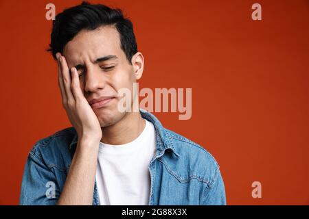 Triste uomo ispanico in camicia accigliato mentre copre il viso isolato su sfondo rosso Foto Stock