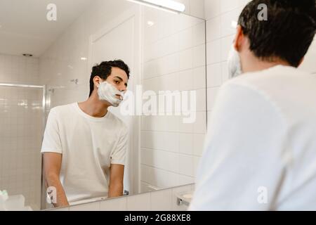 Giovane uomo ispanico che si rade il viso in piedi allo specchio all'interno Foto Stock