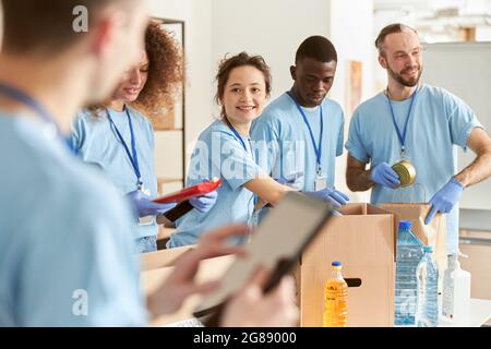 Volontari allegri e diversi sorridono mentre si smistano alimenti in scatole di cartone, lavorando insieme a un progetto di donazione. Focus sulla donna Foto Stock