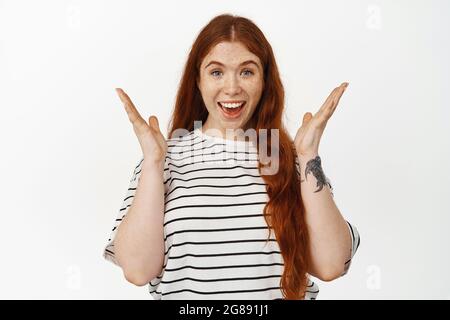 Immagine di sorpresa rossa ragazza sentire grandi notizie, rallegrarsi con positivo e felice espressione del volto, sorridente stupito, eccitato per vedere grande premio, in piedi Foto Stock