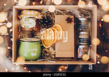 Lviv, Ucraina - 1 dicembre 2017: Marmellata fatta in casa, tè, tazza, spezie, coni di pino sulla paglia in confezione regalo in luci festive. Elegante confezione regalo per il tè per il cristma Foto Stock