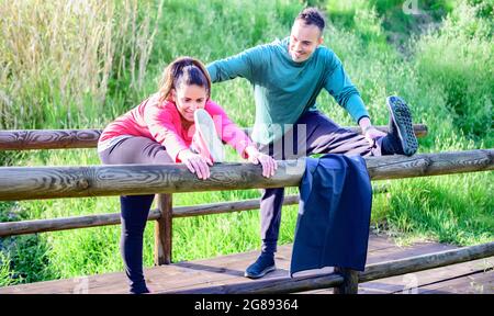 Donna incinta flessibile che si allunga all'aperto con il suo personal trainer - Coppia in abbigliamento sportivo che allunga le gambe e sorride su un parco - Focus on t Foto Stock