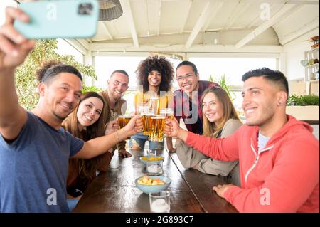 Amici multirazziali che prendono selfie mentre tostano la birra al ristorante - giovani che si divertono con le bevande - concetto di stile di vita su ragazzi e ragazze mak Foto Stock