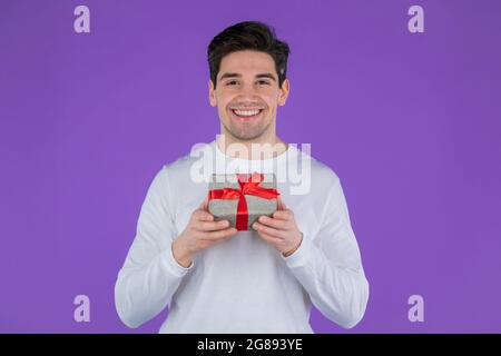 Bell'uomo che tiene una confezione regalo sullo sfondo viola dello studio e sorride alla macchina fotografica. Felice europeo ragazzo ricevuto presente e interessato a ciò che è dentro. Foto Stock