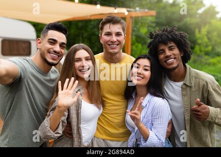 Giovani amici multietnici che prendono selfie insieme vicino a casa del motore, facendo gesti diversi e sorridendo alla macchina fotografica Foto Stock