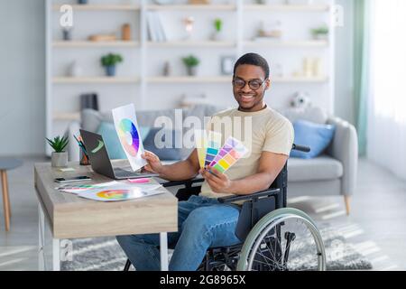Disabled black guy in sedia a rotelle che lavora come graphic designer online, tenendo color Swatch palette, utilizzando computer portatile indoor Foto Stock
