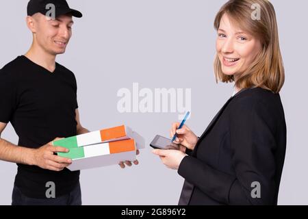 Il corriere in una divisa nera con un cappuccio di picco consegna due scatole per una donna sorridente e lei firma per esso. Foto Stock