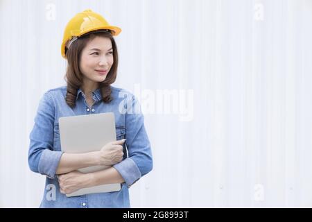 Bella donna asiatica ingegnere in jeans vestito con sicurezza gialla elmetto usando il notebook del computer portatile che fa il lavoro in costruzione sito esterno a. Foto Stock