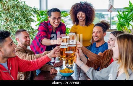 gruppo di amici che festeggiano l'happy hour facendo un brindisi con una pinta di birre all'interno di un bar ristorante. persone multietniche insieme in pub Foto Stock