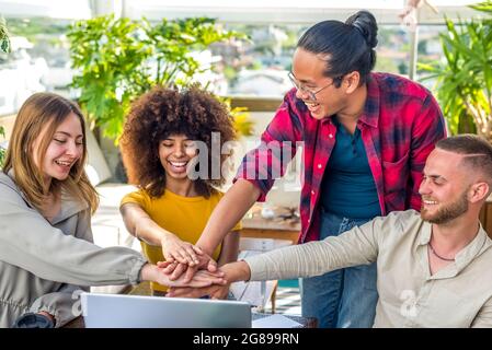 gruppo eterogeneo di amici che festeggiano con un basso cinque la fine di un progetto di startup al coperto seduto su una scrivania da tavolo. studenti multietnici persone insieme Foto Stock