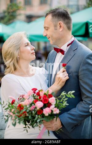 Data romantica in città. Primo piano verticale di coppia matura pronta per la cerimonia di nozze, abbracciandosi l'un l'altro. Blonde donna mette su un fiore boutonnière sulla giacca dello sposo Foto Stock