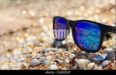 Occhiali da sole con lenti blu si trovano su una spiaggia di conchiglia sabbiosa sul fronte mare in tempo soleggiato. Foto Stock