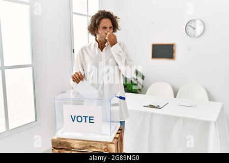 Giovane uomo ispanico votando mettendo envelop in una scatola di voto odore qualcosa di stinoso e disgustoso, odore intollerabile, trattenendo il respiro con le dita su no Foto Stock