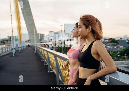 Due corridori asiatici in piedi al ponte che pende contro il bordo del ponte. Le giovani donne si rilassano e guardano all'alba e al tramonto mentre si prendono la pausa dalla corsa Foto Stock