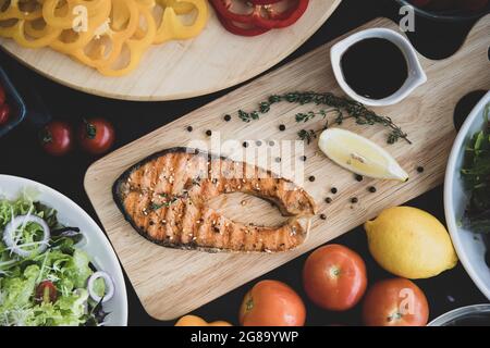 Pezzo di deliziosa bistecca di salmone affettata su un piatto di legno con una varietà di verdure fresche, limone, pomodoro e insalata, ricco di olio di omega 3 e nurri Foto Stock