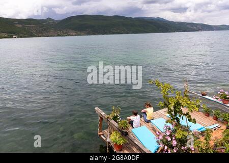 Madre e figlio seduti da sedie a sdraio in legno su un molo sulle rive del tranquillo lago Ohrid, Lin, Albania Foto Stock
