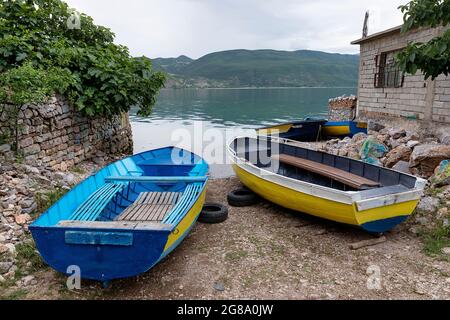 Tradizionali barche da pesca albanesi blu e giallo ormeggiate sulla riva del lago Ohrid dalle case nel villaggio di Lin, Albania Foto Stock