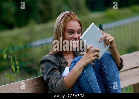 Giovane adolescente bionda fissando alla sua tavoletta in stupore con gli occhi larghi e un grin gleful come lei si rilassa all'aperto su una panchina del giardino Foto Stock