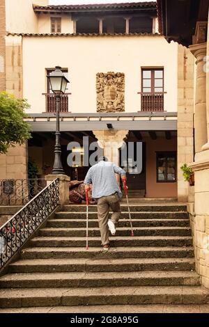 Un uomo con una gamba rotta in un getto sale le scale utilizzando stampelle. Un uomo usa le stampelle per camminare nella città vecchia. Viaggi per disabili. Foto Stock