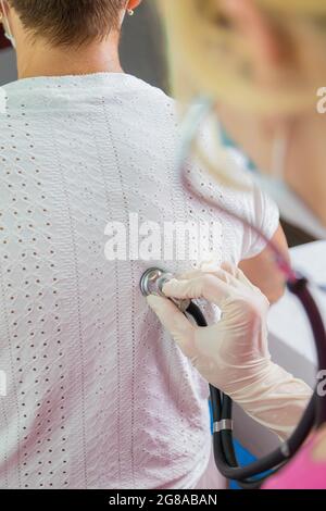 Una ragazza medico in una maschera ascolta con uno stetoscopio alla parte posteriore di una donna adulta. Una donna nell'ufficio del medico si lamenta di una tosse Foto Stock