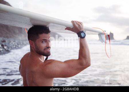 Attraente surfista sorride con la sua tavola da surf poggiata sulla testa, è in spiaggia. Uomo caucasico sportivo e muscolare Foto Stock