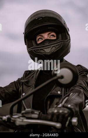 Donna motociclista elegante in casco e giacca in pelle seduta su moto nera. Driver femminile all'aperto su sfondo naturale. Viaggi, caffè racers Foto Stock