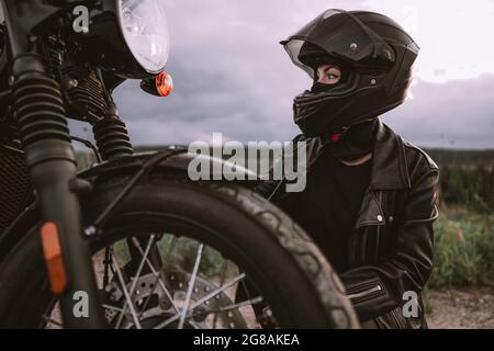 Ritratto di donna motociclista sicura in casco seduto vicino alla bicicletta. Giovane autista che si trova all'aperto al tramonto. Foto Stock