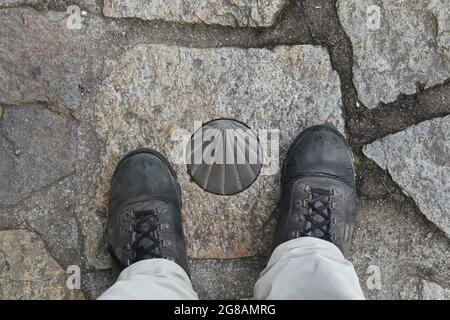 Camino de Santiago (Via di San Giacomo). Stivali da pellegrino raffigurati accanto al tradizionale segno di direzione in bronzo del Camino de Santiago sul pavimento acciottolato a Santiago de Compostela in Galizia, Spagna. La conchiglia del cuoio capelluto, conosciuta anche come conchiglia di San Giacomo, è raffigurata nel segno come un simbolo tradizionale del pellegrinaggio a Santiago de Compostela. Foto Stock