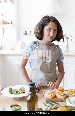 Brunette teen fa panini nella cucina domestica. Bella ragazza sta cucinando nella cucina domestica. Ritratto attraente ragazza in grembiule su un ki chiaro Foto Stock