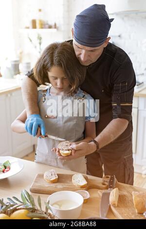 Uno chef genitore insegna a cucinare a sua figlia. Padre e figlia stanno cucinando insieme nella cucina domestica. La cucina dei sandwich italiani. Scatto verticale. Foto Stock