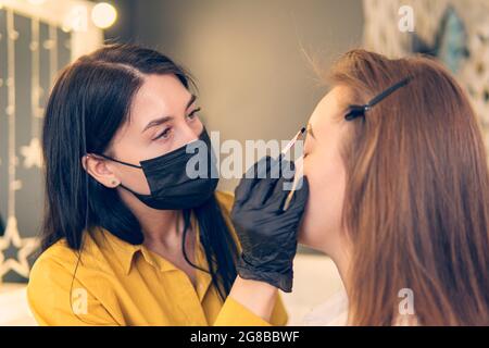 procedura di tintura delle sopracciglia, la ragazza asciuga la vernice dalle sue sopracciglia. Un cosmetologo maestro dipinge le sopracciglia di un cliente nel primo piano del salone. Foto Stock