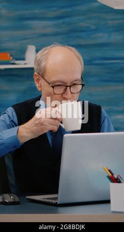 Rapporti di lettura dell'uomo d'affari senior seduti davanti al laptop che lavora da casa che beve il caffè. Uomo in pensione che utilizza la tecnologia moderna analizzando la ricerca della digitazione mentre la moglie si siede sul divano leggendo un libro Foto Stock
