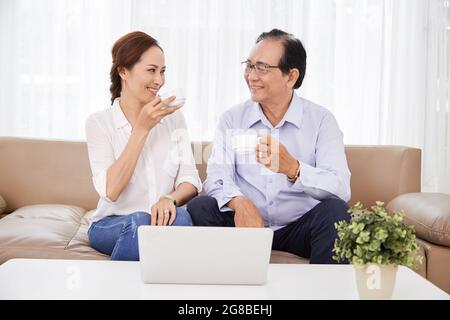 Felice uomo anziano e donna che beve caffè e si guarda l'un l'altro quando si siede sul divano con il computer portatile aperto Foto Stock