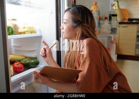 Donna pensiva con i generi alimentari di controllo del notepad nel suo frigorifero e scrivendo giù una lista dei prodotti che deve comprare Foto Stock