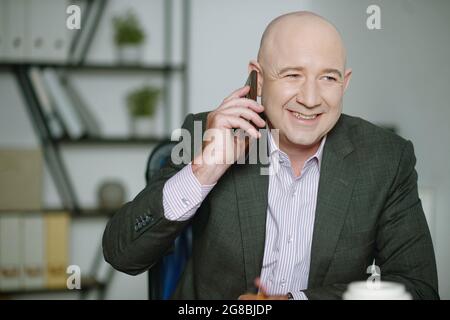 Uomo d'affari maturo di successo seduto al tavolo in ufficio e parlando sul telefono cellulare con sorriso Foto Stock