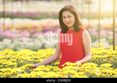 Carina donna asiatica in abito rosso in piedi in giardino fiorito con molti tipi e fiori colorati. Bella luce del sole sullo sfondo. Foto Stock