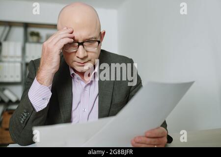 Uomo d'affari maturo ponderato in occhiali seduto al tavolo in ufficio e l'esame del contratto di affari Foto Stock