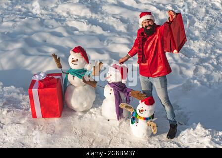 Uomo eccitato con simpatico pupazzo di neve in elegante cappello e sciarpa su campo innevato. Buon inverno famiglia pupazzo di neve con regalo. Madre neve-donna, padre neve-uomo e. Foto Stock