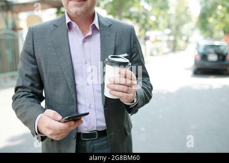Primo piano di uomo d'affari in tuta con telefono cellulare e caffè bevendo mentre si cammina in città Foto Stock