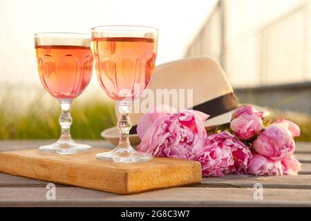 due bicchieri con vino rosato e bouquet di pony rosa. deliziosa bevanda rinfrescante, fiori enormi e cappello da uomo sul molo in legno al tramonto Foto Stock