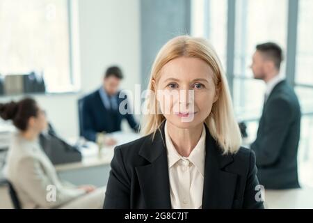 Ritratto di una donna d'affari bionda matura che guarda la macchina fotografica mentre i suoi colleghi si siedono alla riunione all'ufficio Foto Stock