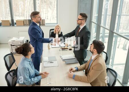 Due partner commerciali che si levano in piedi e scuotono le mani durante la riunione di affari, concludono un accordo fra loro Foto Stock