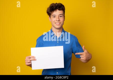 Bellissimo scolaro europeo in camicia blu con carta bianca vuota su sfondo giallo. Foto Stock