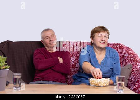 Un uomo vecchio che dorme su un divano, la sua moglie che mangia popcorn e guarda il televisore con due bicchieri d'acqua a un tavolo. Foto Stock