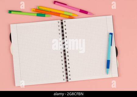 Aprire il quaderno quadrato della scuola e le penne colorate sullo sfondo rosa. Foglio bianco vuoto di libro sulla tabella. Materiali di consumo per ufficio sul desktop Foto Stock