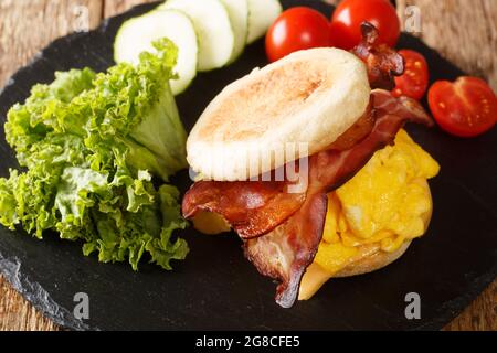 Primo piano di un sandwich per la colazione con uova strapazzate, pancetta e formaggio su un muffin inglese su una tavola di ardesia sul tavolo. Orizzontale Foto Stock