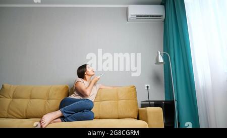 Donna giovane che regola il condizionatore d'aria mentre si siede sul divano. Foto Stock
