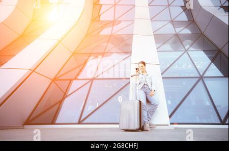 Giovane donna con valigia da viaggio con telefono cellulare alle porte dell'aeroporto. Acquisto online dei biglietti aerei e richiesta di prenotazione rapida degli hotel Foto Stock