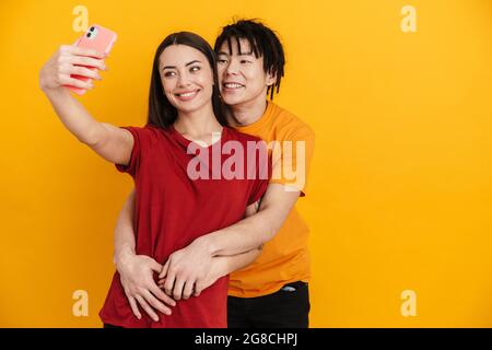 Felice giovane coppia multietnica su sfondo giallo, prendendo un selfie, abbracciando Foto Stock